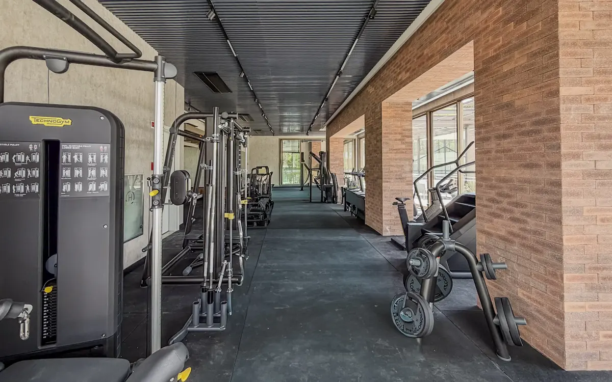 Academia do Florà Alphaville com aparelhos de musculação, bicicleta, escada ergométrica e anilhas sobre piso emborrachado escuro, ao lado de parede de tijolinho com aberturas para o jardim