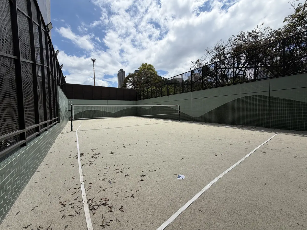 Quadra de beach tennis do Florà Alphaville com piso de areia clara, rede, linhas de demarcação brancas, paredes revestidas de pastilhas verdes com desenho de montanhas e fechamento em tela metálica