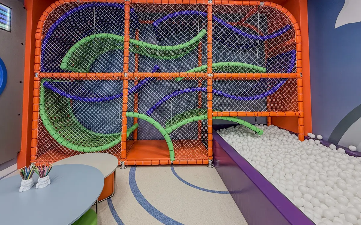 Playground da brinquedoteca do Florà Alphaville com rede laranja e tubos coloridos para escalar, piscina de bolinhas brancas, mesa baixa com lápis de cor e parede azul-escura