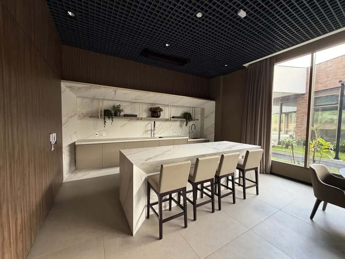 Espaço gourmet do Florà Alphaville com bancada de porcelanato em formato de ilha e quatro banquetas estofadas, copa com prateleiras douradas e plantas, painéis de madeira e janela para o jardim