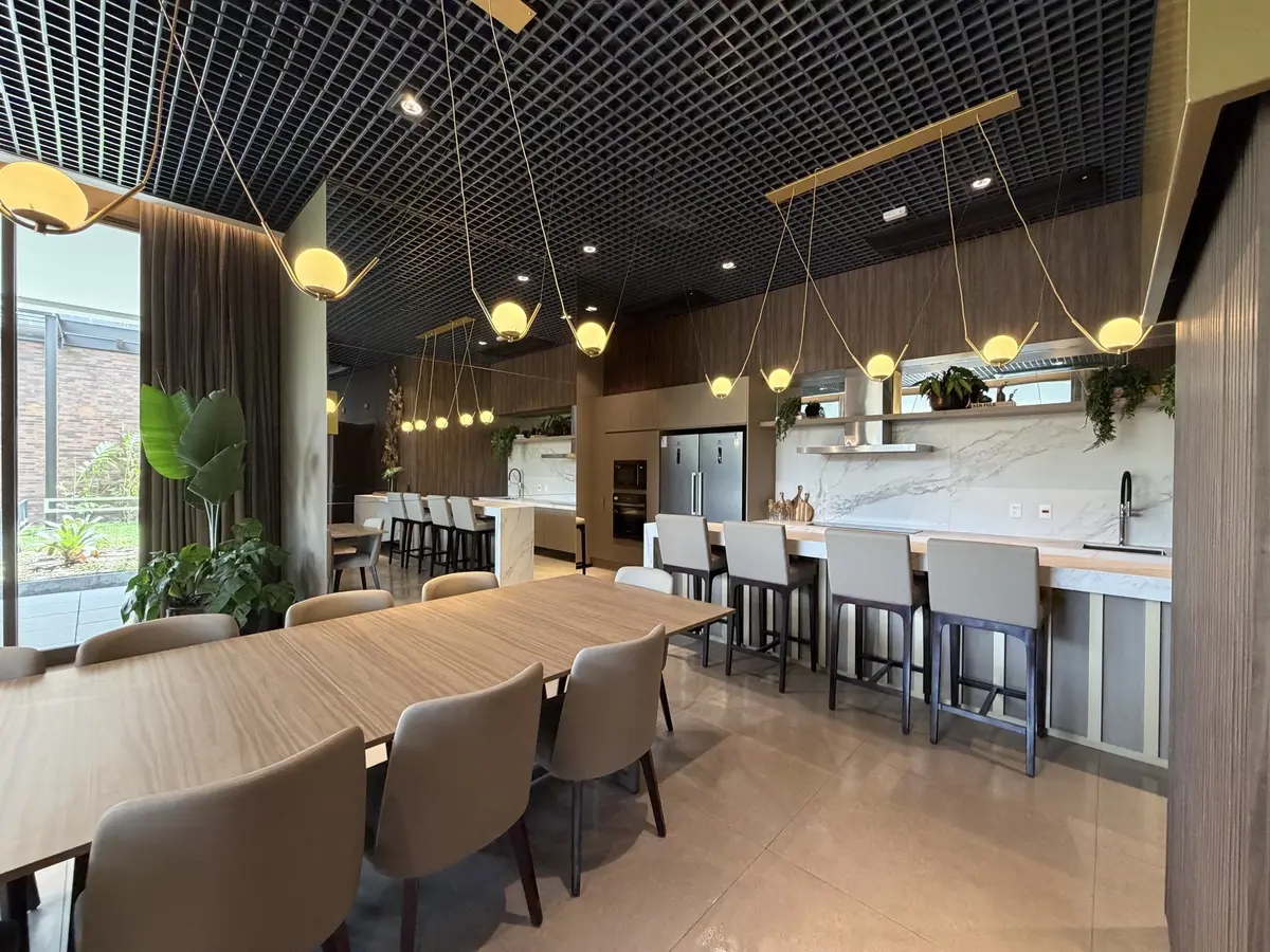 Espaço gourmet do Florà Alphaville com mesa de madeira e cadeiras estofadas, bancada com banquetas, cooktop, forno, geladeira, pendentes de globos dourados e forro de grelha escura