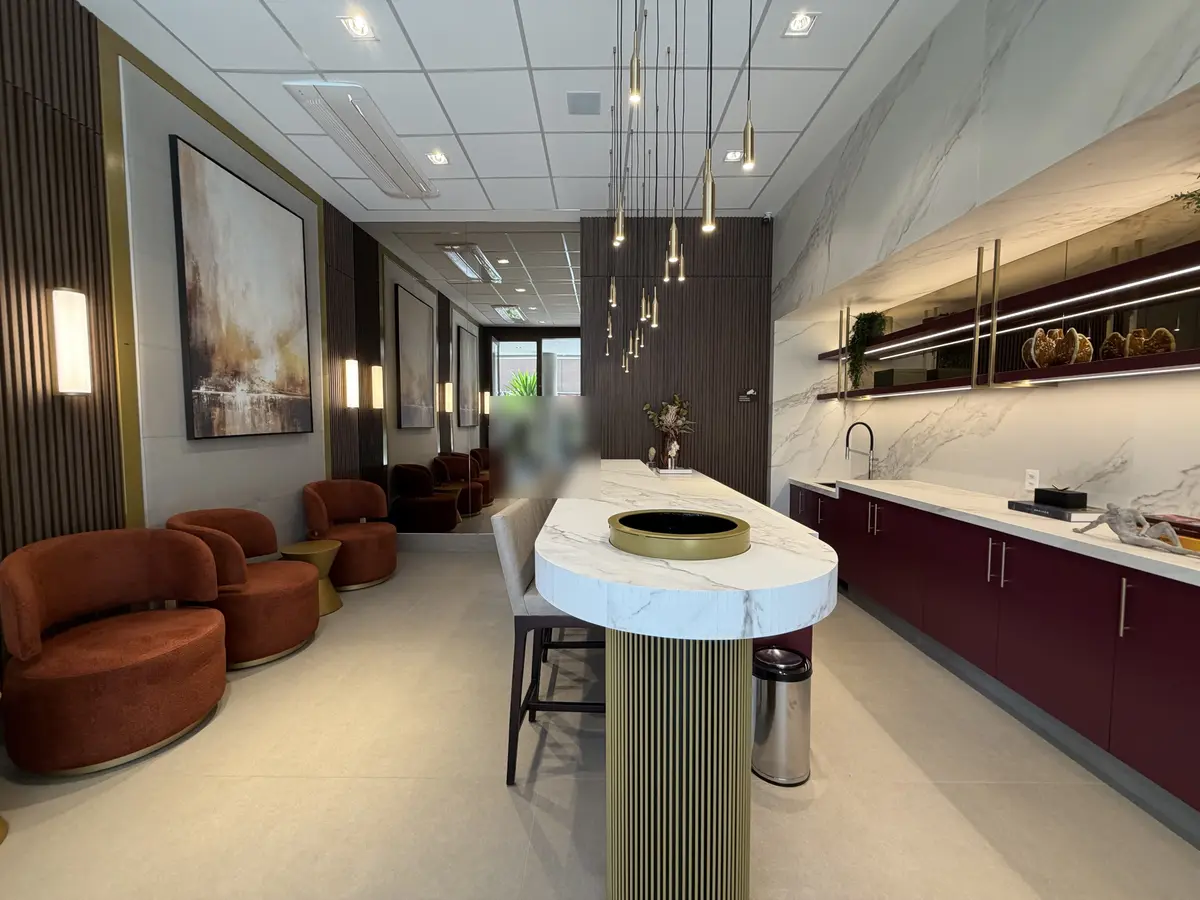 Ambiente social do Florà Alphaville visto do fundo, com poltronas cor de telha, bancada de porcelanato com banquetas, copa com armários bordô e prateleiras iluminadas, pendentes dourados e painéis ripados