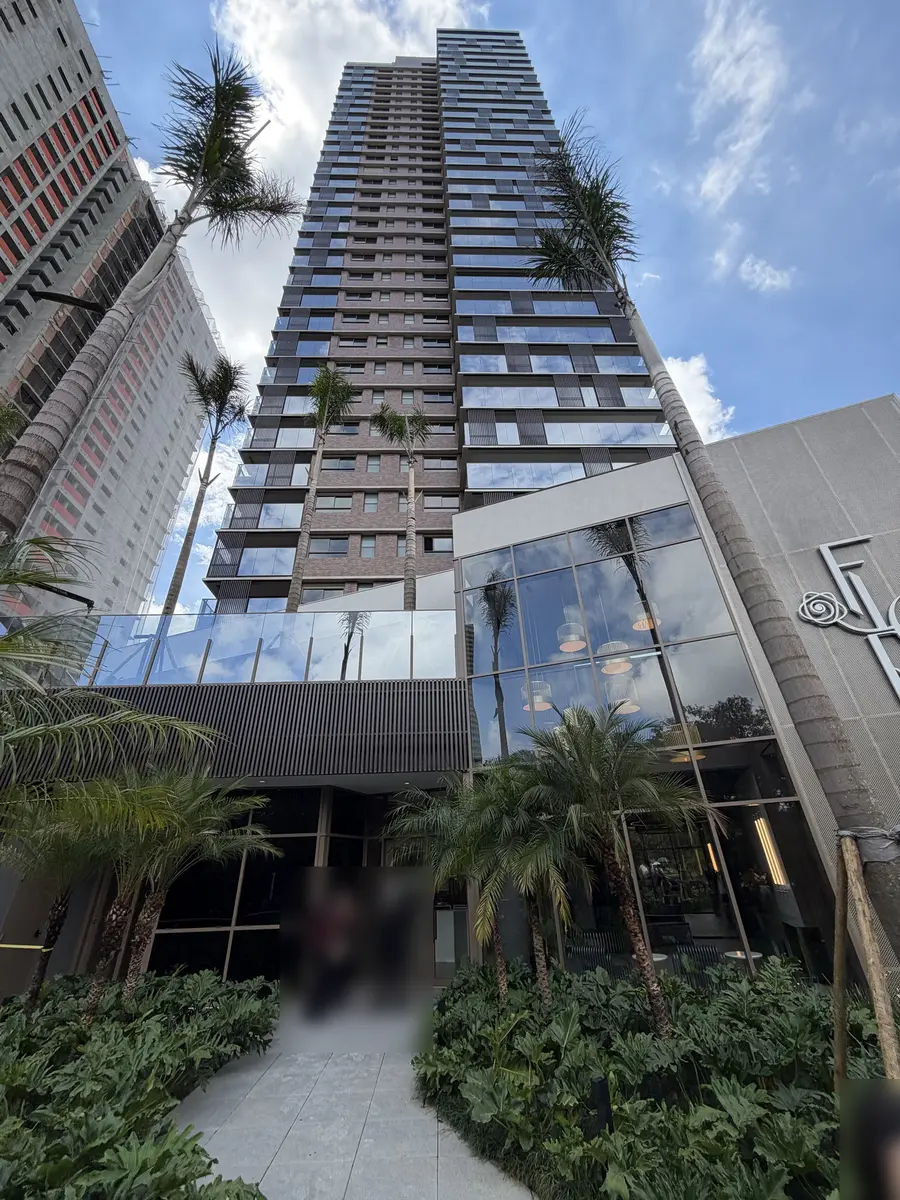 Fachada do Florà Alphaville vista de baixo, com a torre de varandas envidraçadas e tijolinho, palmeiras, entrada com jardim e o logotipo do condomínio com uma rosa
