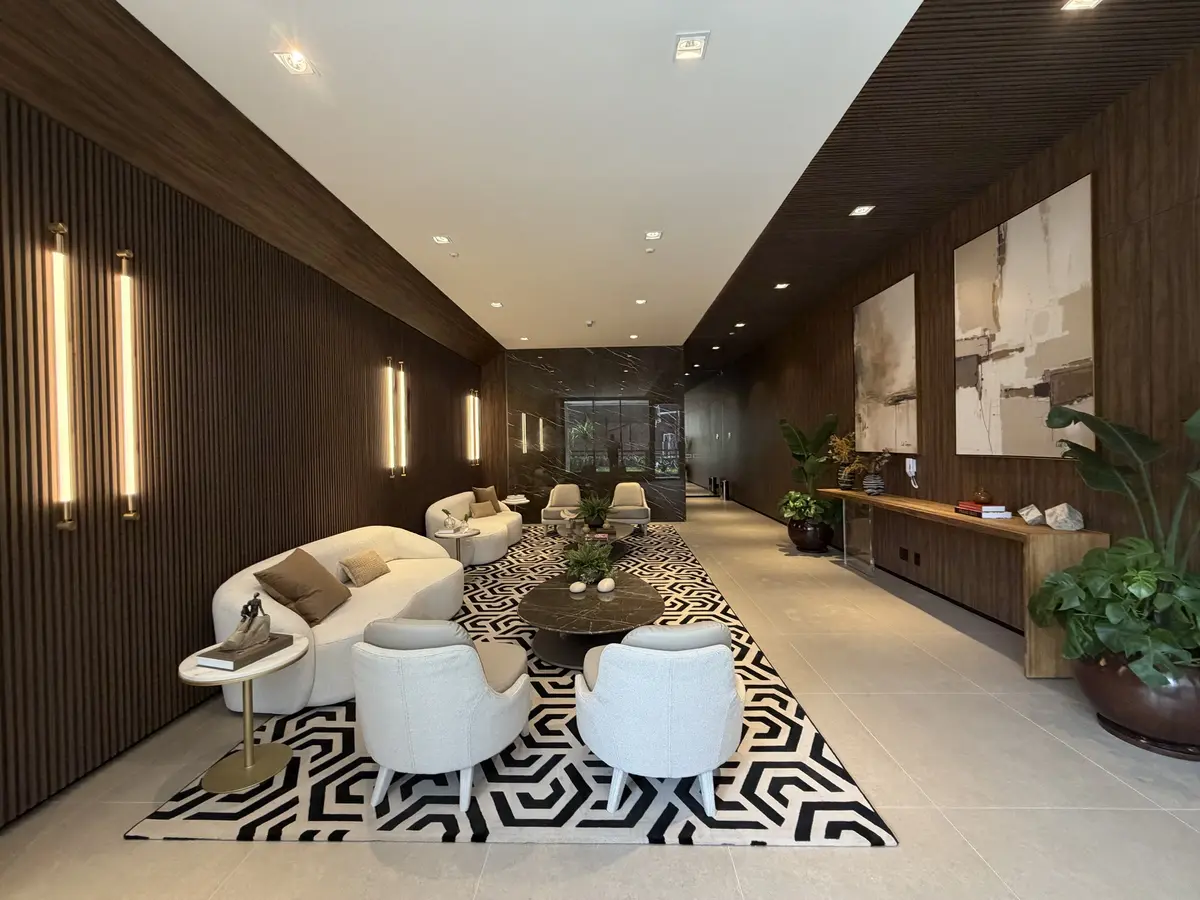 Hall de estar do Florà Alphaville com painéis ripados de madeira, sofás curvos brancos, poltronas, tapete geométrico preto e branco, mesa de centro de mármore escuro e aparador de madeira com vasos de plantas