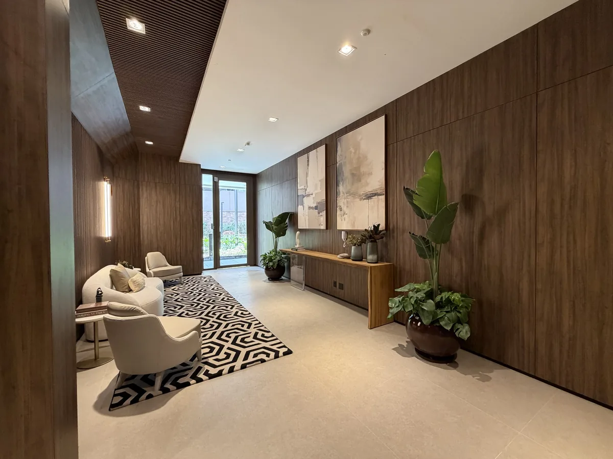 Hall social do Florà Alphaville com painéis de madeira, sofá e poltronas claras, tapete geométrico, quadros e vasos de plantas, com porta de vidro para o jardim
