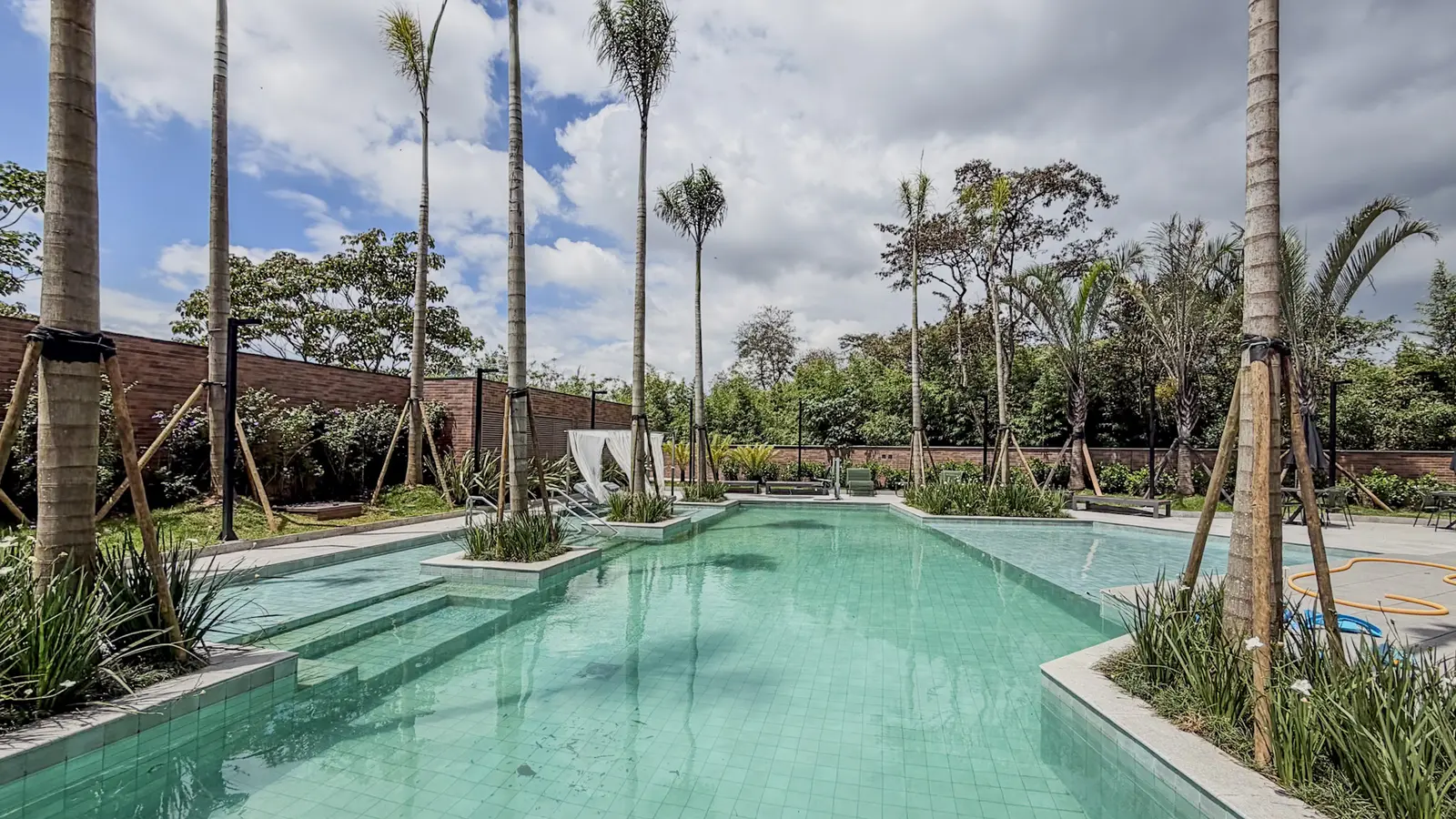 Piscina descoberta do Florà Alphaville com revestimento verde-água, deck de pedra clara, palmeiras jovens e a mata ao fundo, na Alphagran, em Barueri