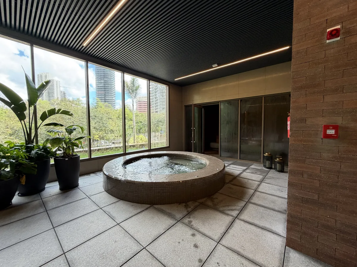 Hidromassagem circular com revestimento em pastilhas no spa do Florà Alphaville, ao lado de janelas amplas com vista para as árvores e vasos de plantas