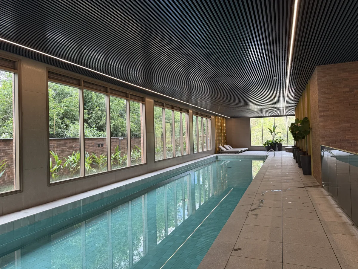 Piscina coberta do Florà Alphaville vista em ângulo, com revestimento verde-azulado, janelas para o jardim, parede de tijolinho, vasos de plantas e espreguiçadeiras ao fundo