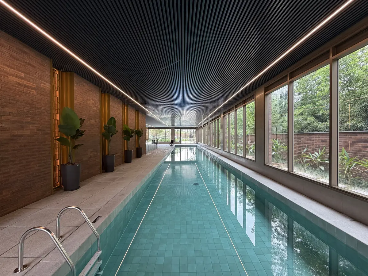 Piscina coberta do Florà Alphaville vista de frente, com linhas de luz no fundo, parede de tijolinho com vasos de plantas à esquerda, janelas para o jardim à direita e forro ripado escuro