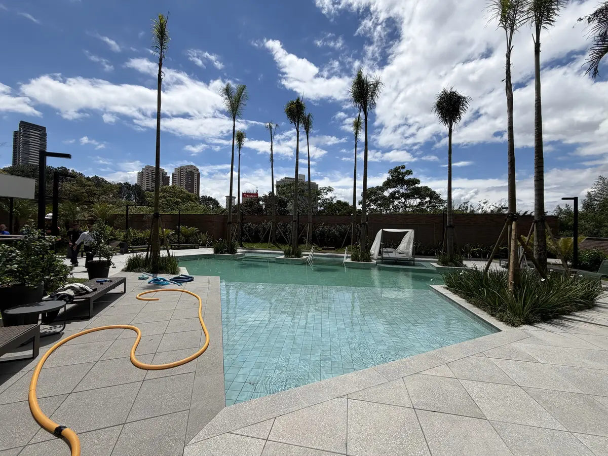 Piscina descoberta do Florà Alphaville com revestimento verde-claro, deck de pedra clara, palmeiras jovens, cama de descanso com cortinas brancas e espreguiçadeiras junto ao paisagismo