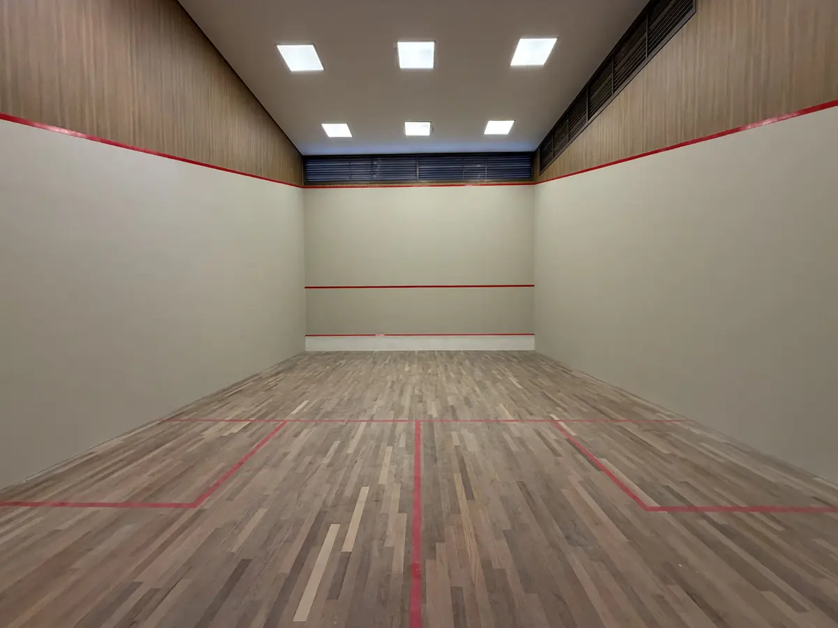 Quadra de squash do Florà Alphaville com piso de madeira, paredes lisas com faixas vermelhas de marcação, forro com luminárias embutidas e venezianas no alto