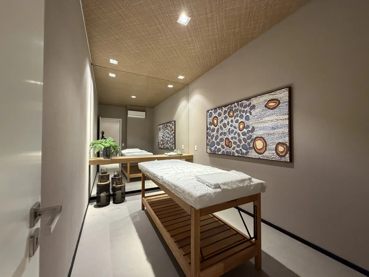 Sala de massagem do Florà Alphaville vista da porta, com maca de madeira, forro texturizado, espelho, ar-condicionado e quadro em tons de azul e laranja