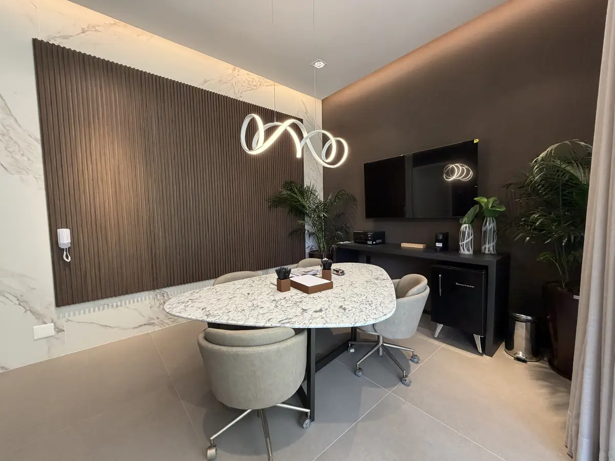 Sala com mesa oval de pedra clara e cadeiras estofadas, painel ripado de madeira sobre parede de porcelanato, pendente luminoso escultural, TV, frigobar retrô e plantas