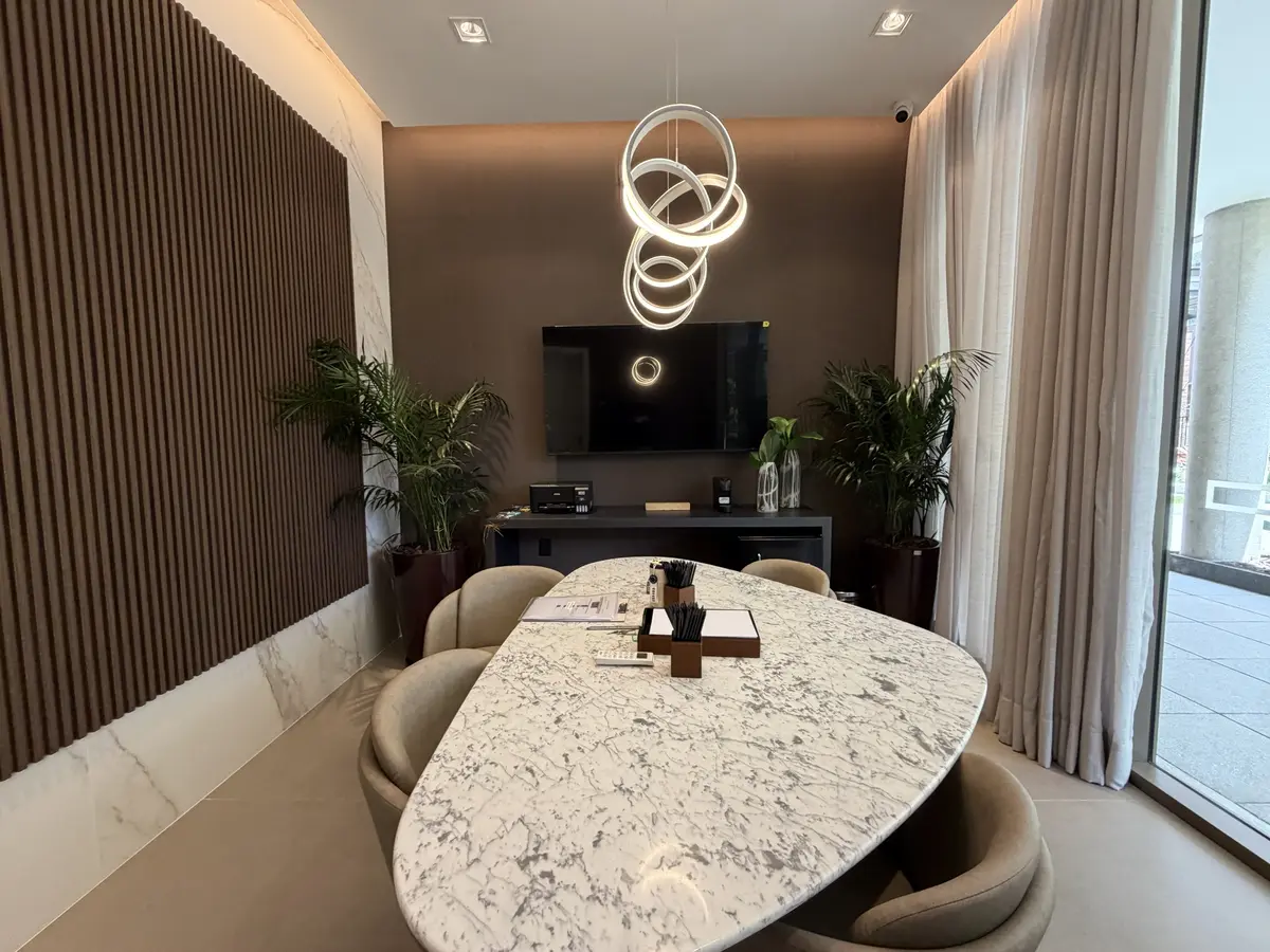 Sala com mesa oval de pedra clara e cadeiras estofadas vista de frente, com TV na parede, pendente de anéis luminosos, painel ripado, cortina de linho e janela para o jardim