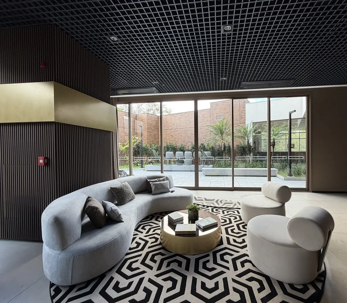 Salão de festas do Florà Alphaville com sofá curvo cinza, poltronas redondas, mesa de centro dourada, tapete geométrico preto e branco e portas de vidro para o jardim