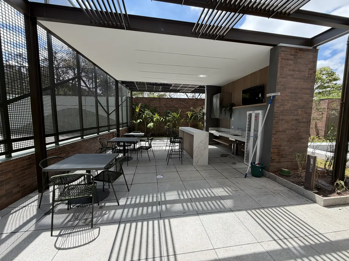 Sport bar do Florà Alphaville em área coberta por pergolado de ripas, com mesas e cadeiras verdes, bancada de porcelanato, TV na parede de madeira e jardim com plantas tropicais